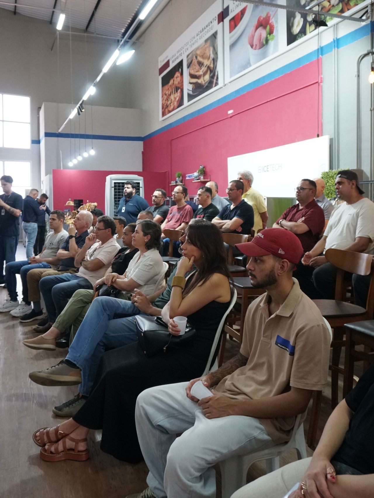 Um grupo de pessoas em uma sala, assistindo a uma apresentação. Muitas estão sentadas, olhando para a frente. Parede rosa e azul.