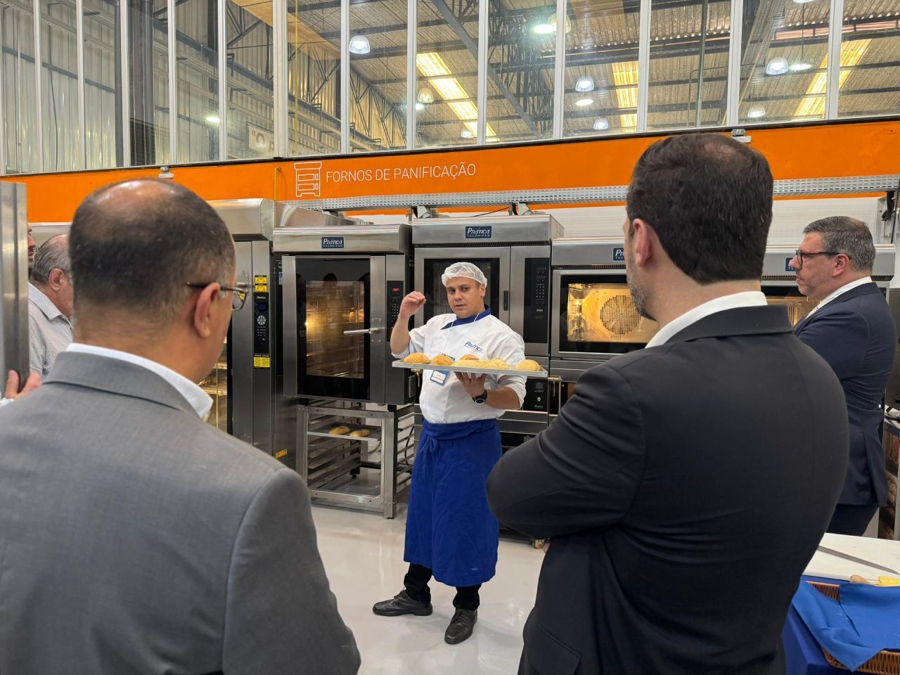 Um chef de avental azul apresentando produtos assados ​​para um grupo em frente aos fornos.