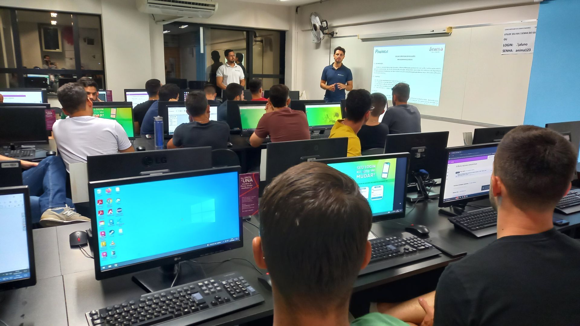 Um grupo de pessoas está sentado em frente a monitores de computador em um laboratório de informática.