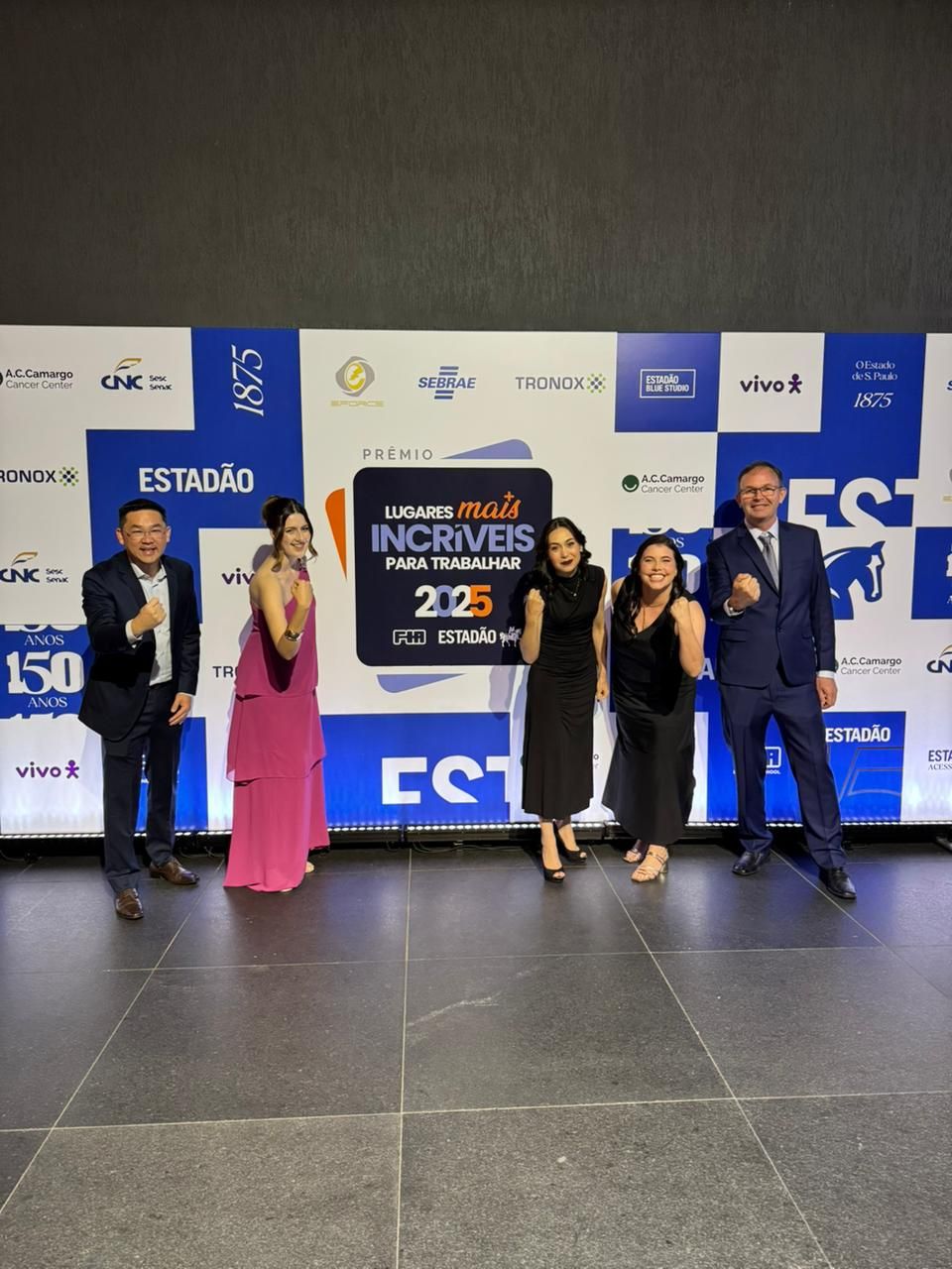 Cinco pessoas posam ao lado de uma faixa, comemorando uma premiação. Elas estão em frente a um fundo azul, sorrindo.