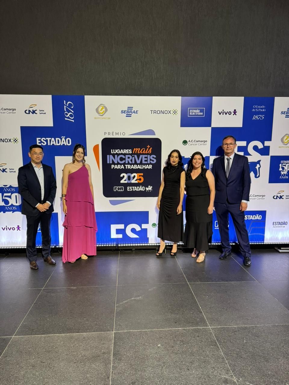 Cinco pessoas estão em pé em frente a um painel com um logotipo. Duas mulheres de vestido, três homens de terno, todos sorrindo.