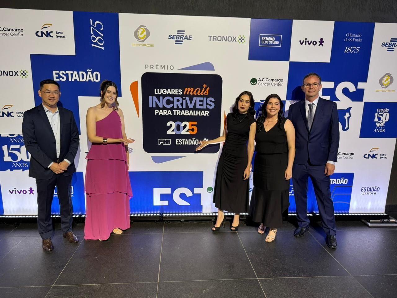 Grupo de cinco pessoas posando em frente a um painel com logotipos e texto. Duas mulheres de vestido, três de terno.
