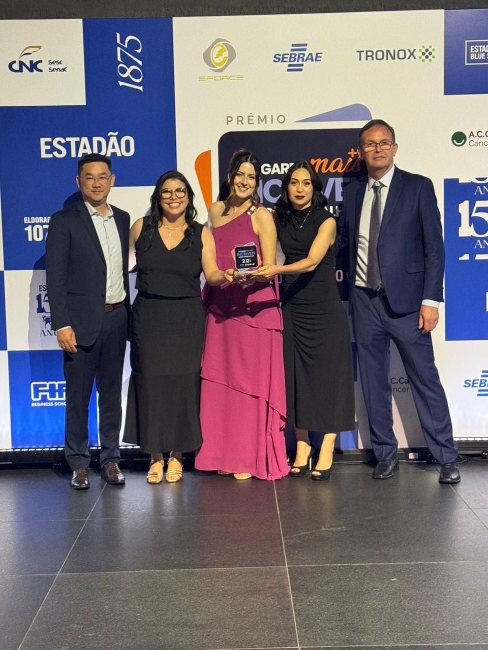 Cinco pessoas segurando um prêmio em um palco com uma faixa.