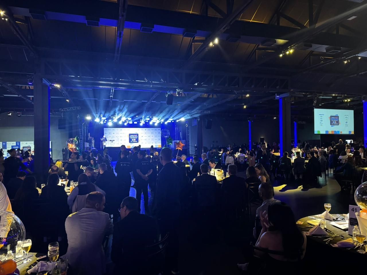 Grande evento em um salão de baile com pouca luz, com pessoas, palco e luzes de palco azuis.