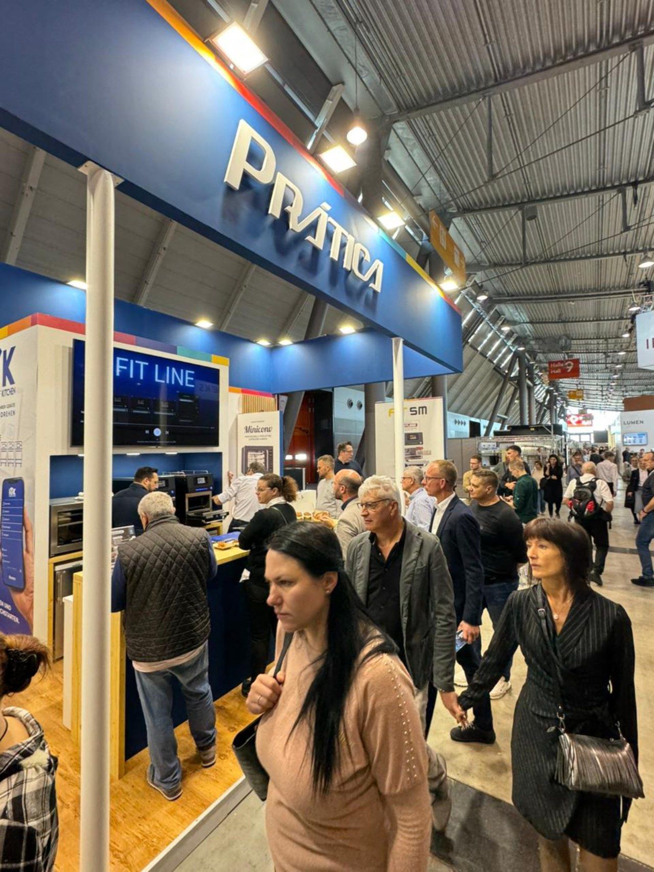 Um grupo de pessoas está caminhando em frente a um estande de pratika