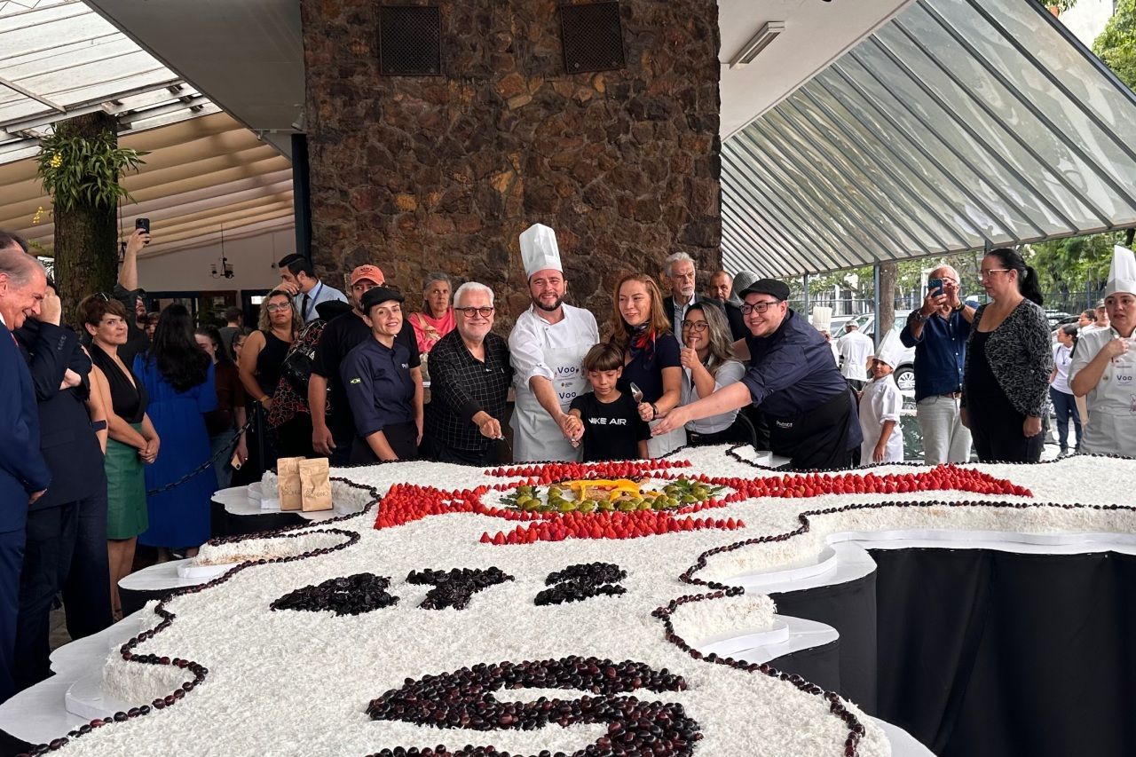 Um grande bolo sendo cortado por chefs e outras pessoas, em frente a um prédio; cerejas e frutas por cima.