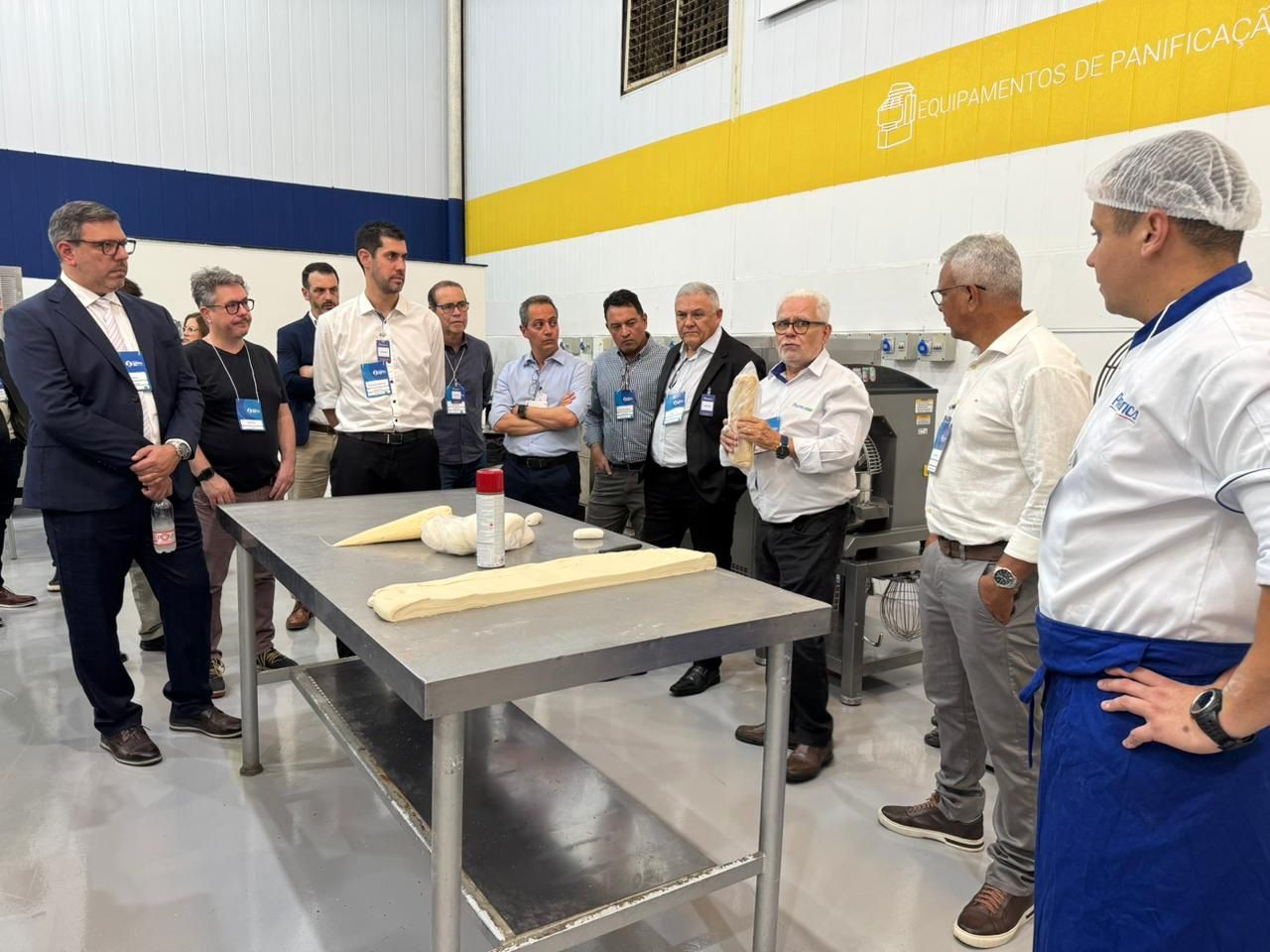 Grupo de homens em uma área de processamento de alimentos observa uma demonstração. Um chef segura uma amostra de pão.