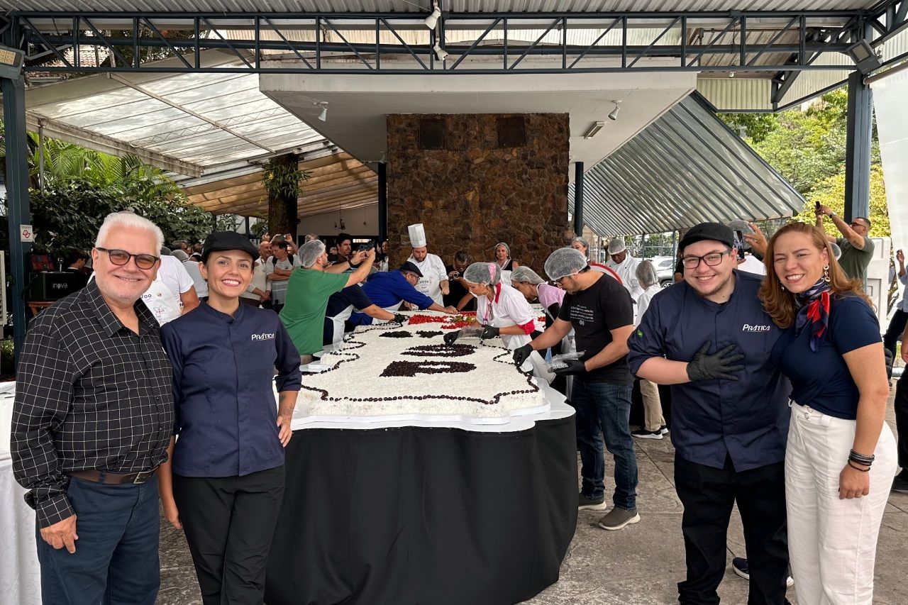 Grupo posa com um bolo gigante, chefs ao fundo. Ambiente externo.