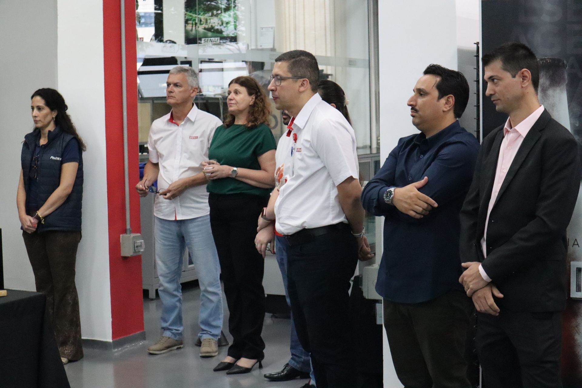Grupo de pessoas em pé dentro de um ambiente fechado, algumas olhando para frente, outras observando. Detalhes em vermelho e branco ao fundo.