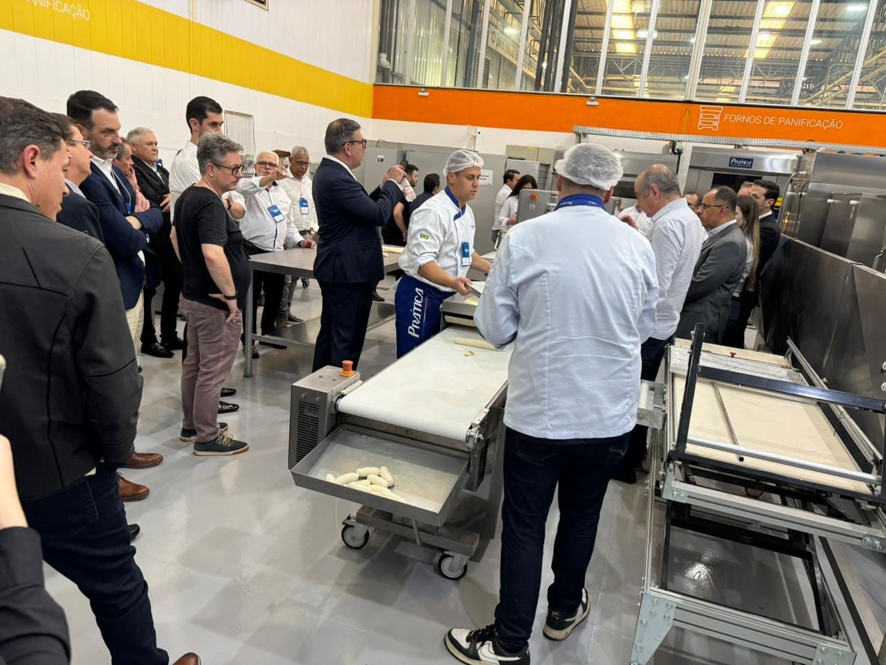 Grupo observa padeiro trabalhando em uma padaria moderna. Uniformes brancos, maquinário e detalhes em amarelo são visíveis.