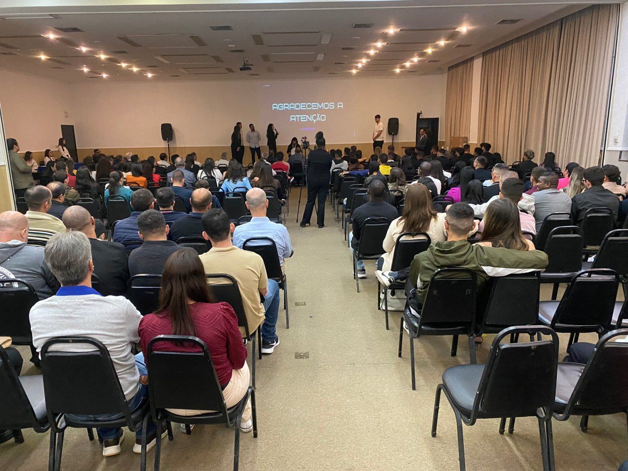 Um grande grupo de pessoas está sentado em cadeiras em uma sala grande.