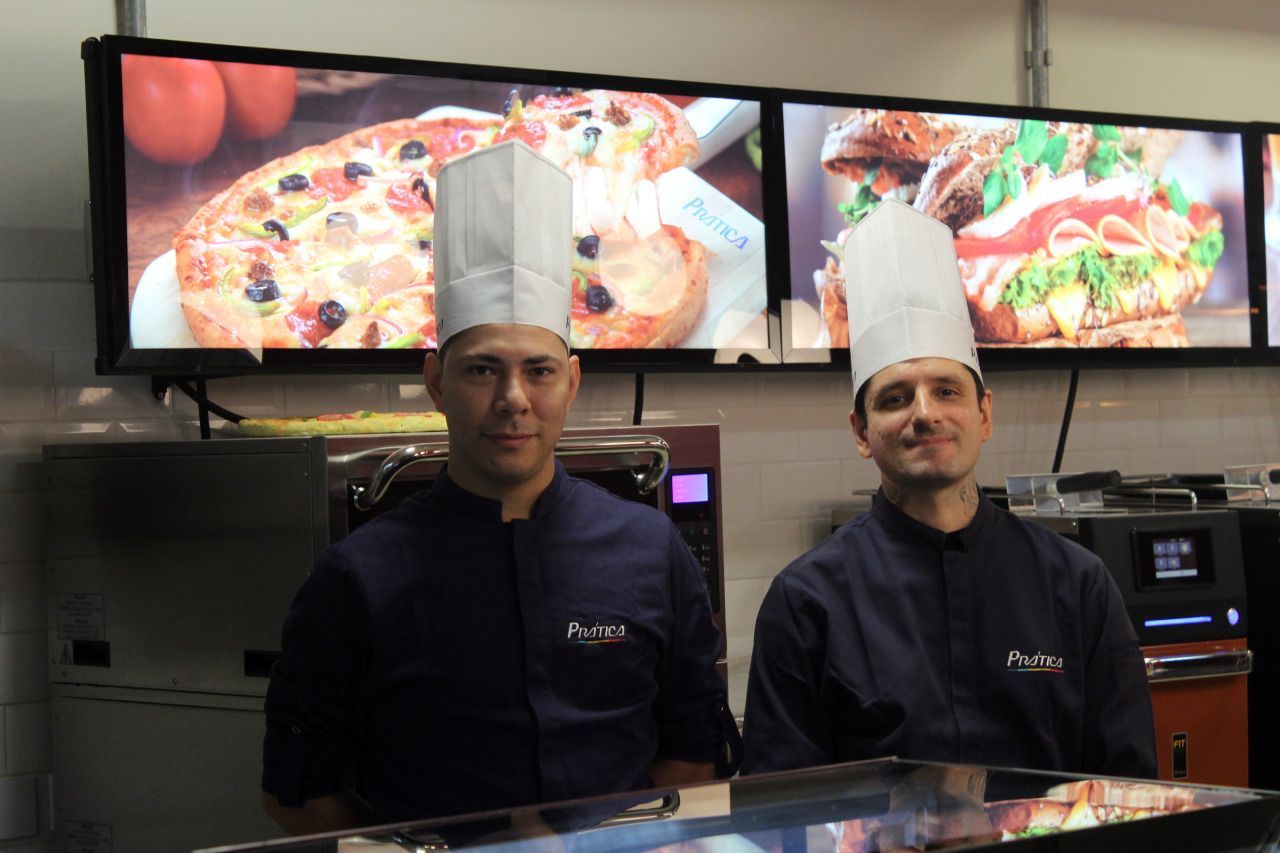 Dois homens usando chapéus de chef estão em frente a uma exposição de pizzas e sanduíches