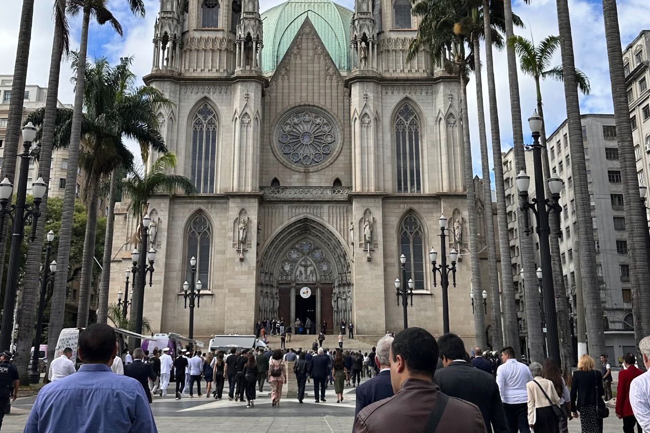 Entrada da catedral com pessoas, ladeada por altas palmeiras.