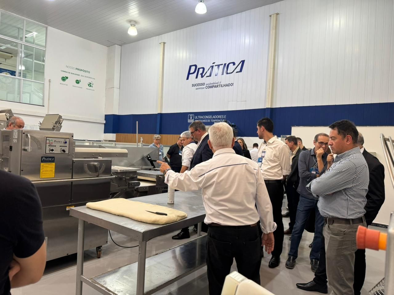 Grupo em uma cozinha comercial observa uma demonstração, com um apresentador apontando para o equipamento.