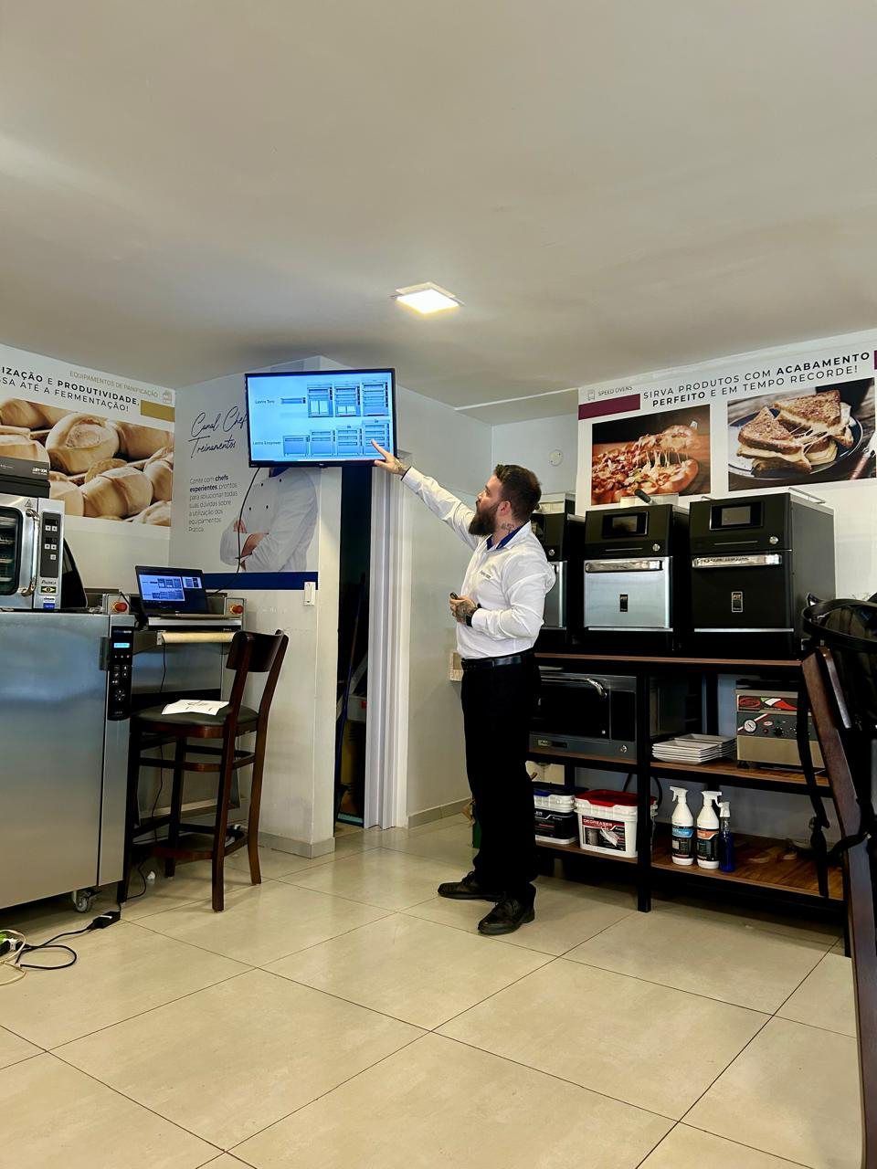 Um homem está de pé na cozinha apontando para uma televisão.