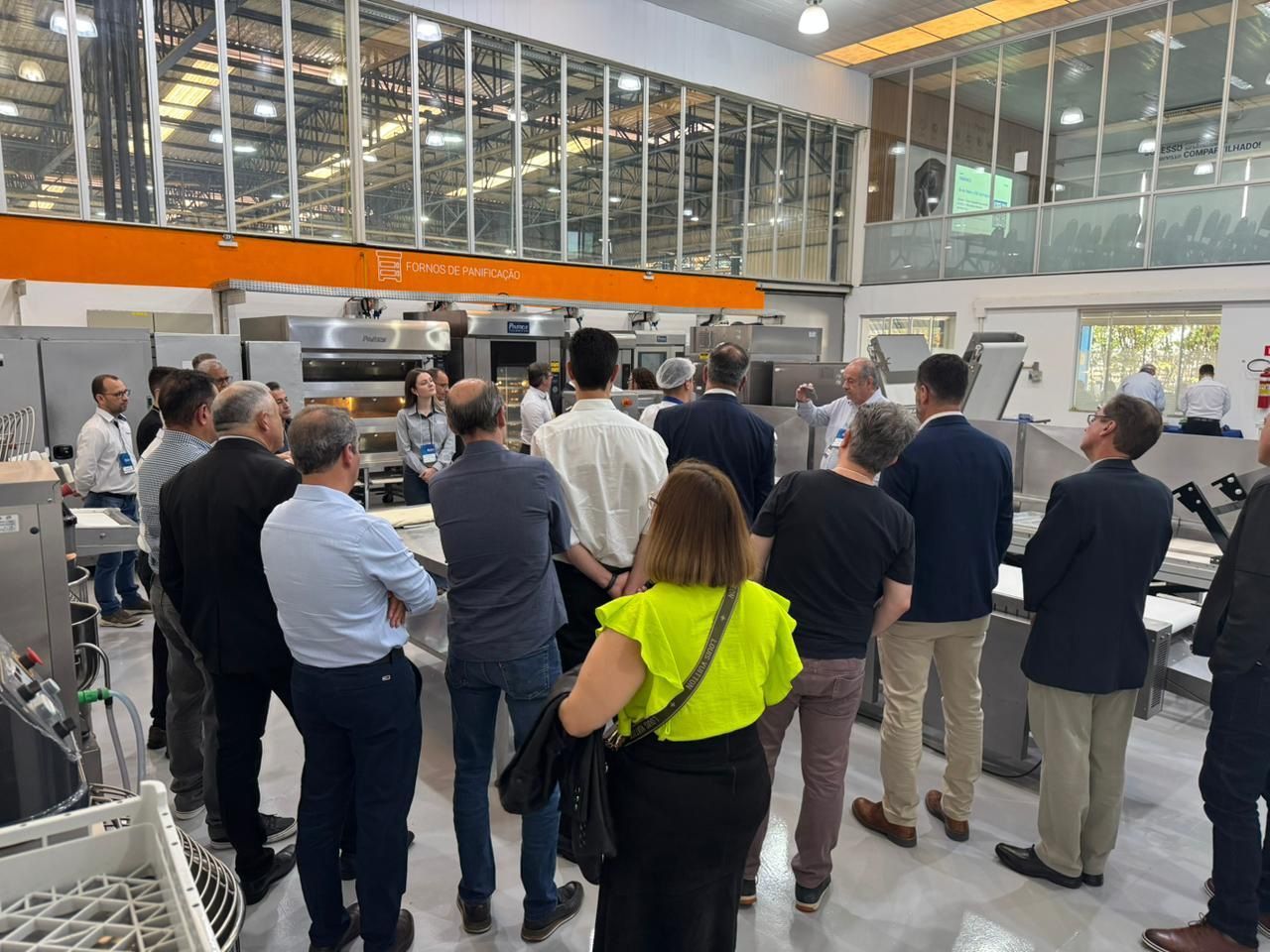 Grupo de pessoas visitando uma padaria, observando fornos e equipamentos. Sala iluminada, ambiente industrial.