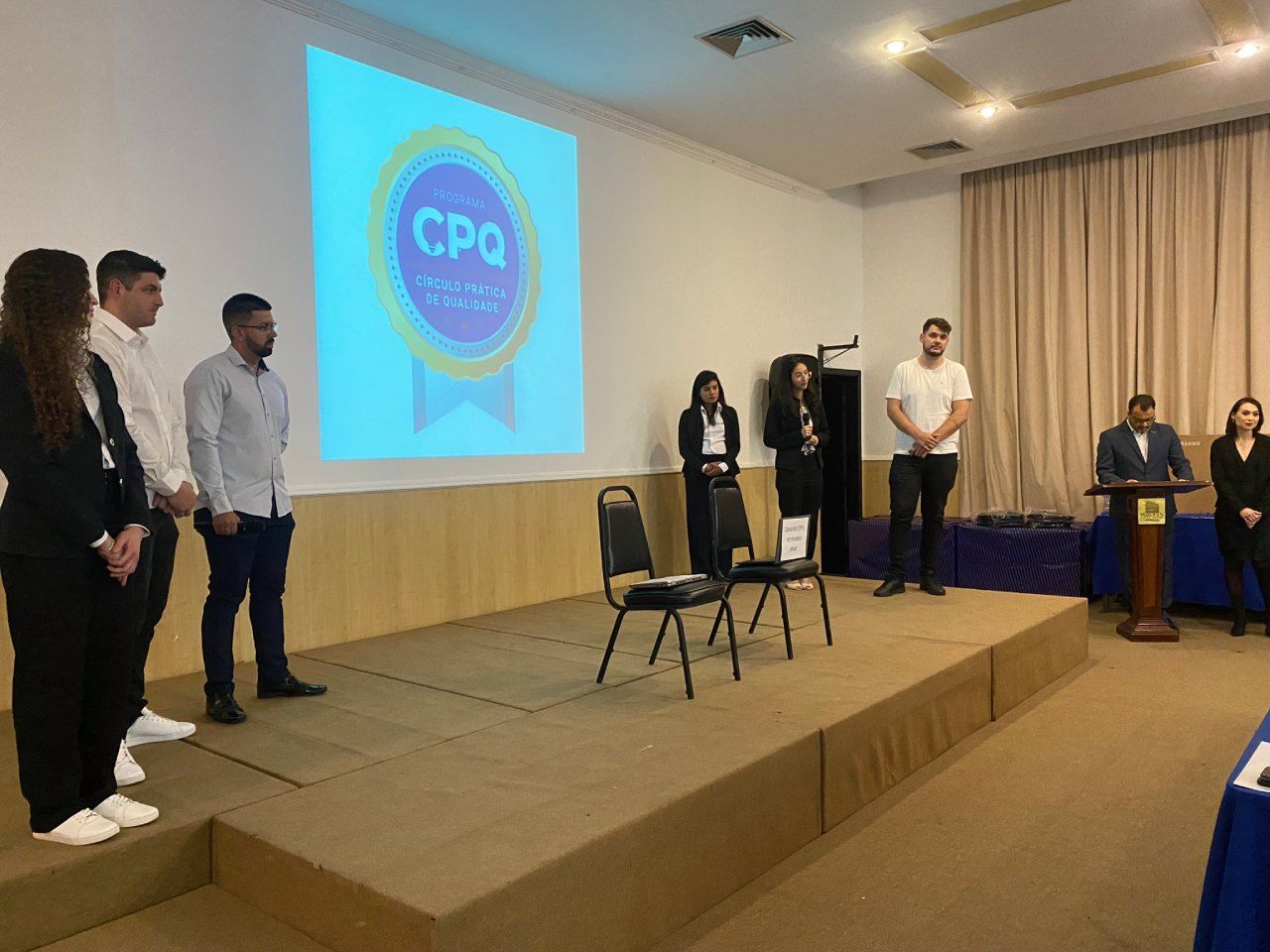 Um grupo de pessoas está em pé em um palco em frente a uma tela grande.