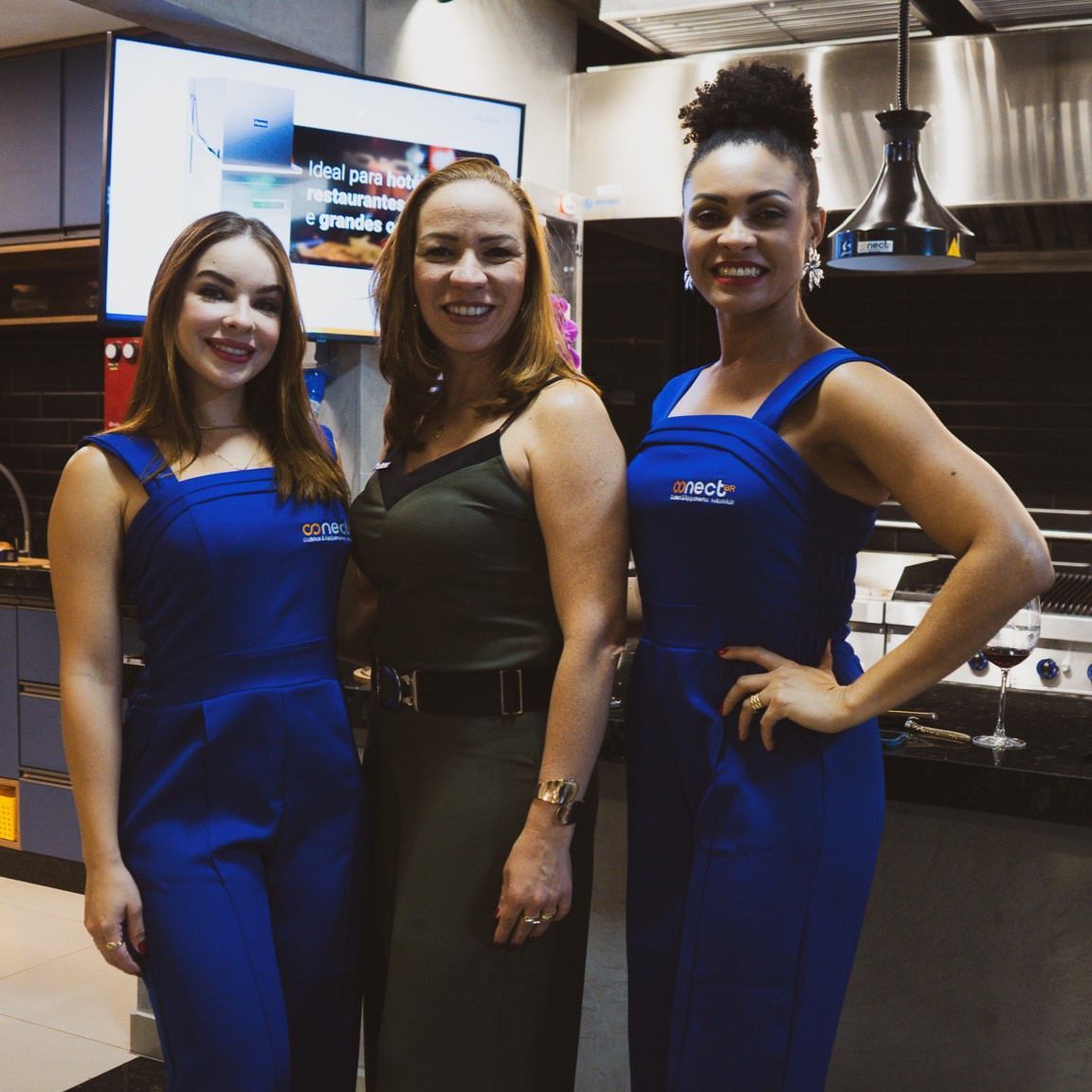 Três mulheres de macacão azul posam para uma foto em uma cozinha
