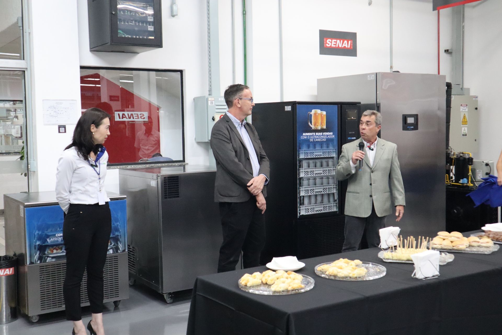 Três pessoas conversando em um evento em uma fábrica, perto de um local onde eram servidos refrescos.