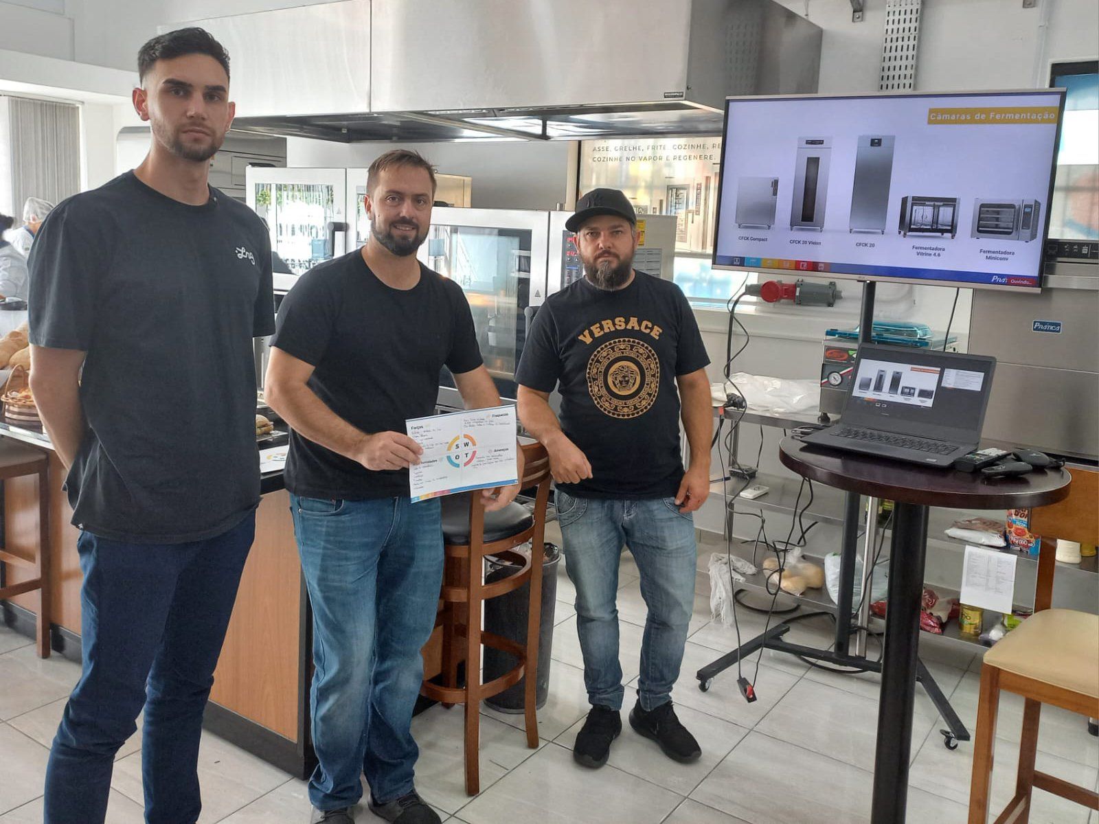 Três homens estão de pé, um ao lado do outro, em uma cozinha, segurando um certificado.