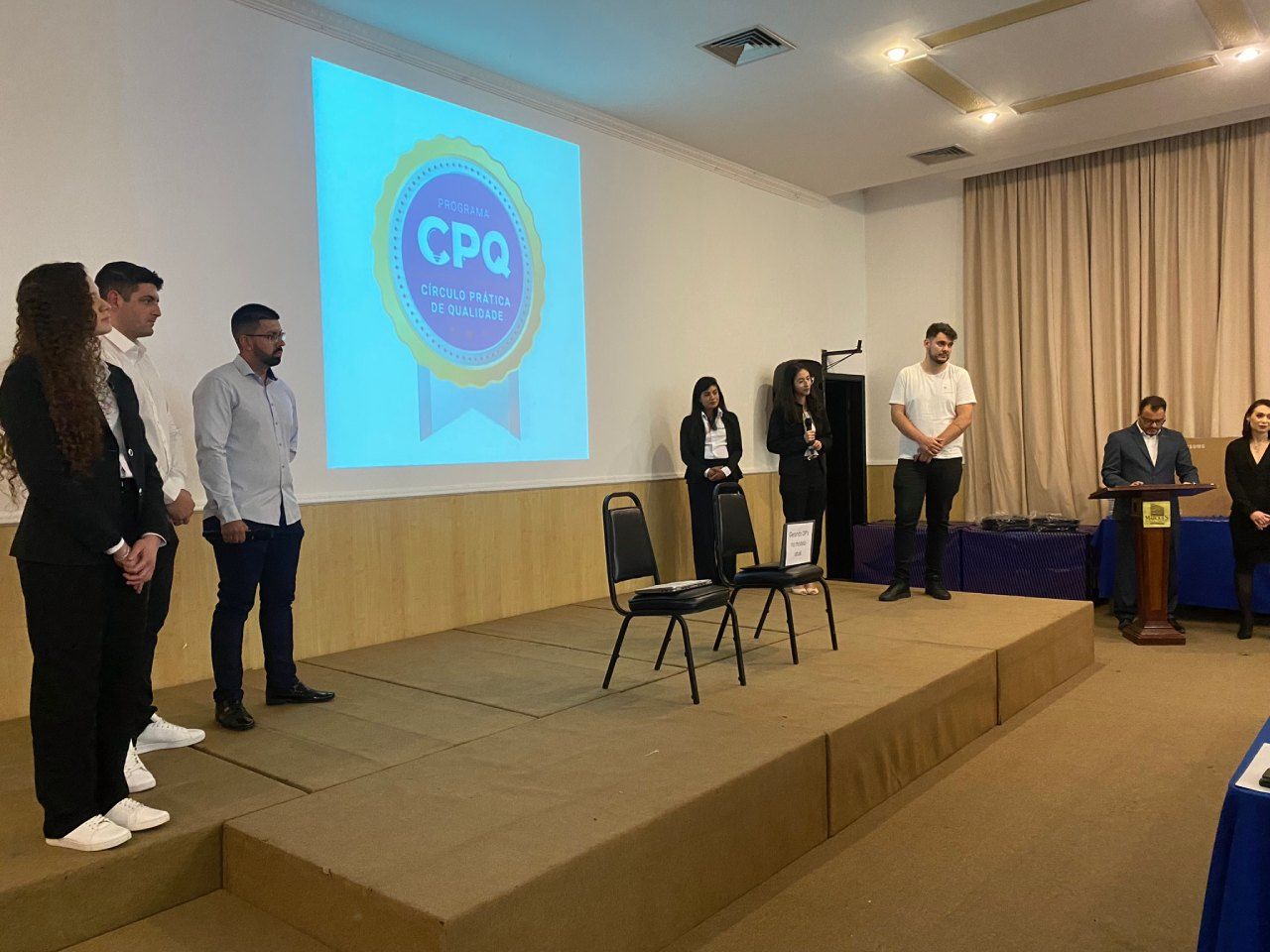 Um grupo de pessoas está em pé em um palco em frente a uma tela de projetor.