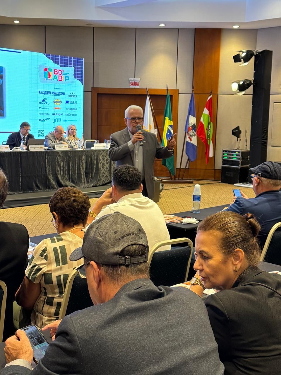 Um homem está falando em um microfone na frente de um grupo de pessoas em uma sala de conferências.