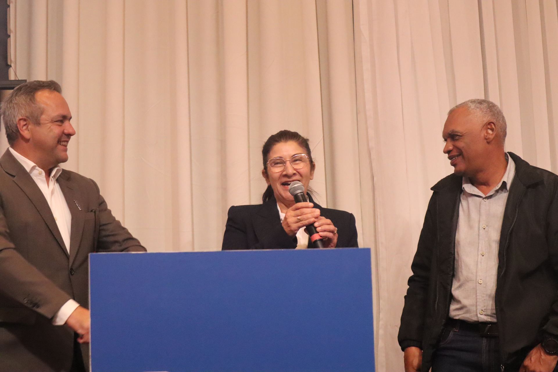 Uma mulher fala em um microfone enquanto dois homens estão atrás dela.