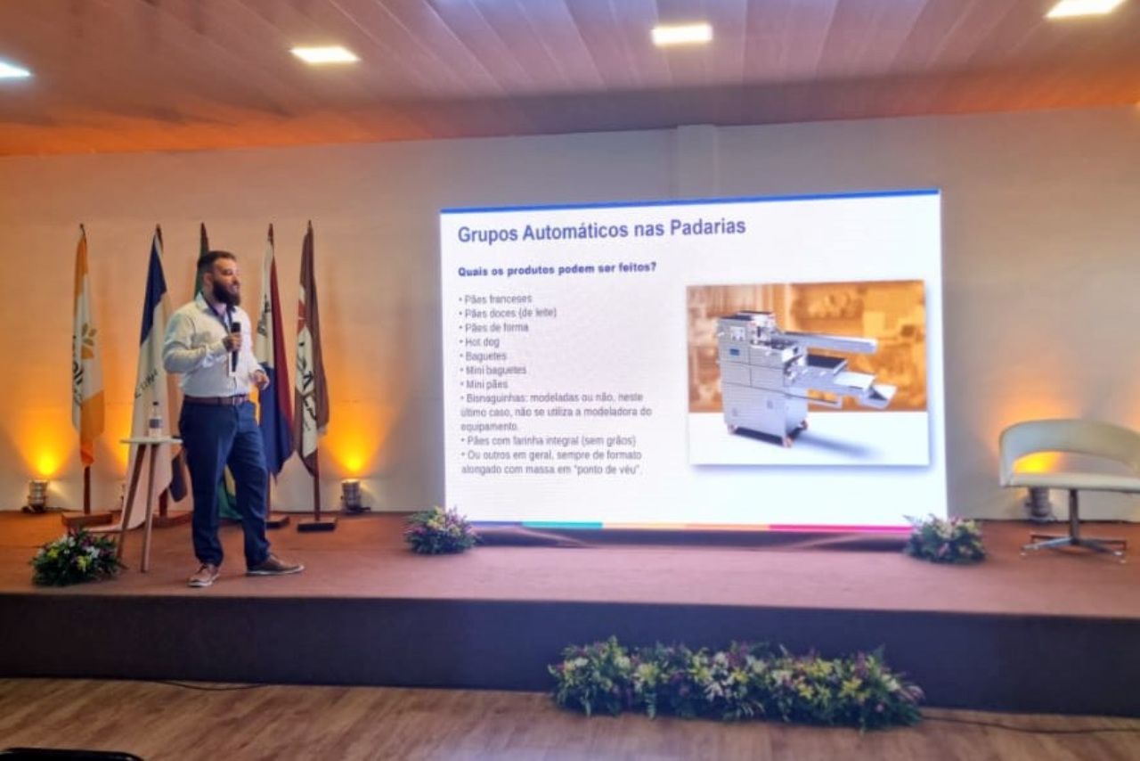 Um homem está fazendo uma apresentação em um palco em frente a uma tela grande.