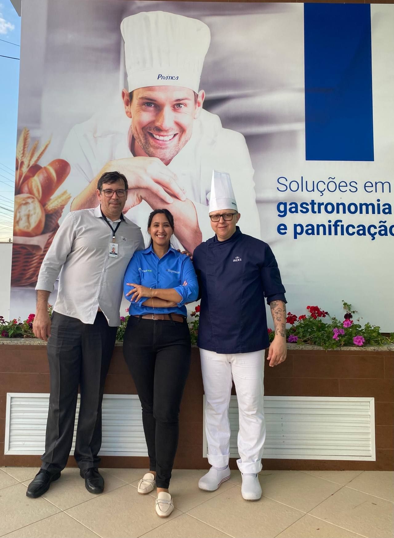 Três pessoas estão posando para uma foto em frente a um pôster de um chef.