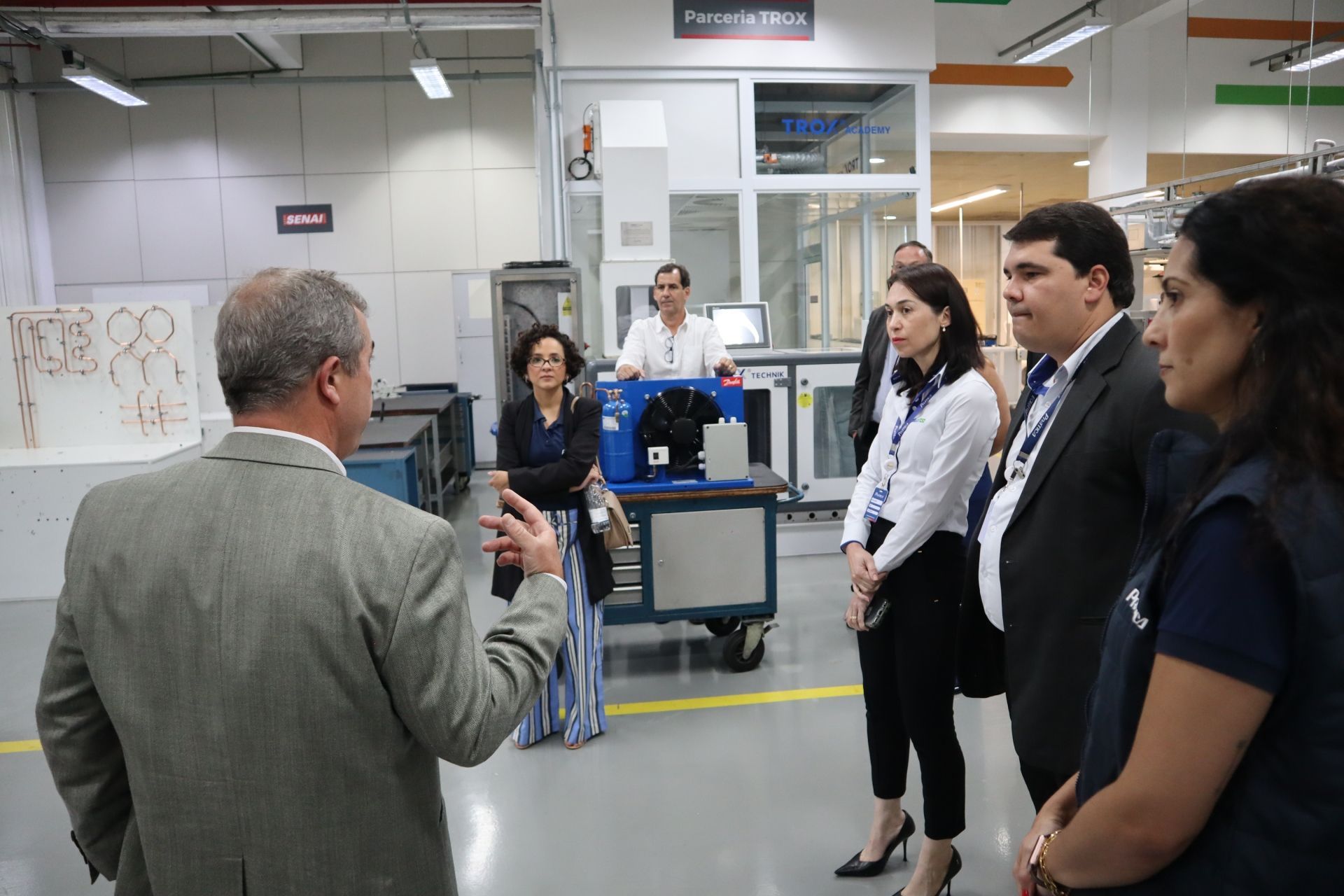 Grupo de pessoas em um ambiente fabril, ouvindo um homem gesticular. Máquinas e equipamentos são visíveis ao fundo.