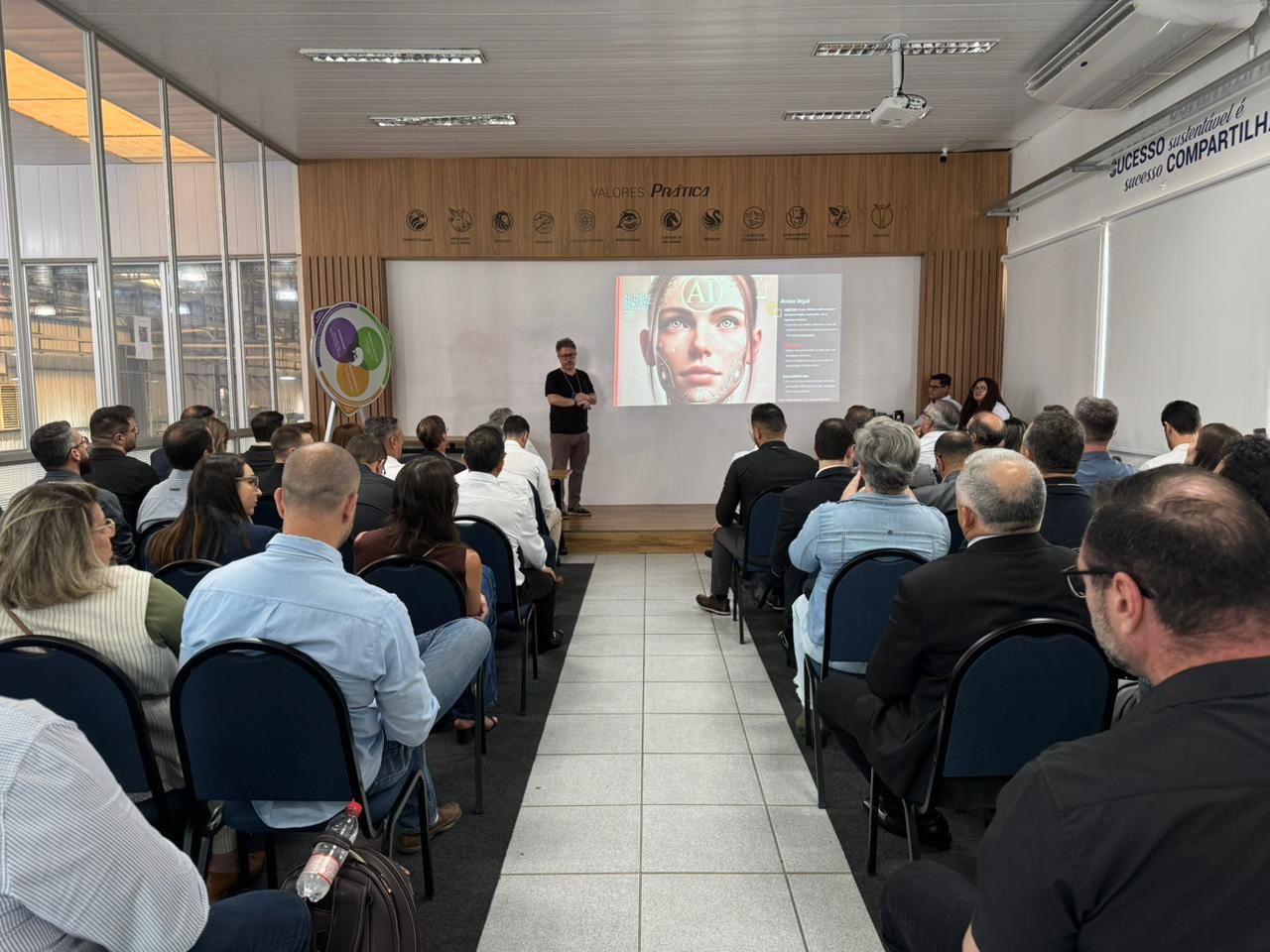 Uma apresentação é feita para uma plateia sentada. Um homem está em pé diante de uma tela com um rosto humano.