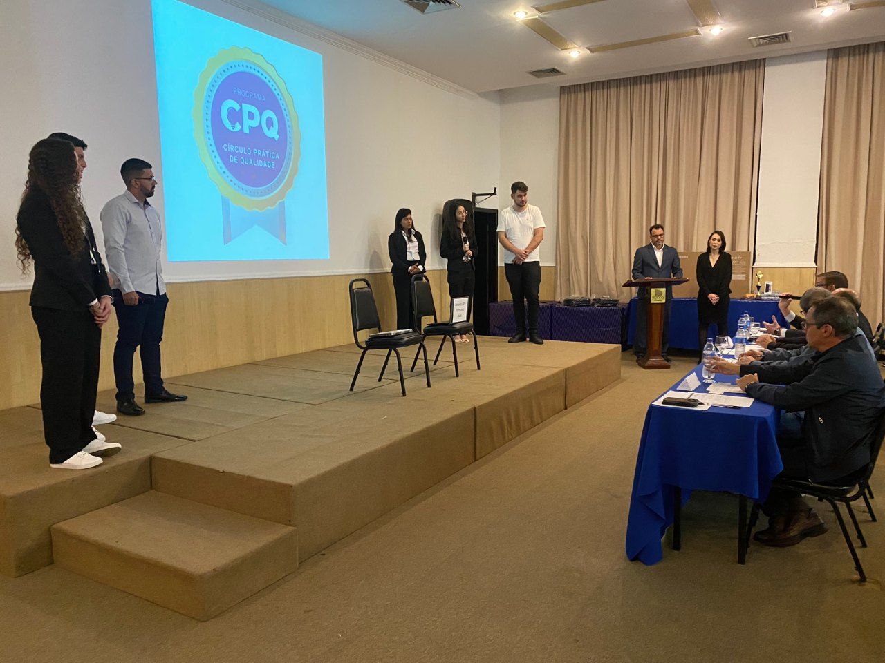 Um grupo de pessoas está em pé em um palco em frente a uma tela de projetor que diz cpq.