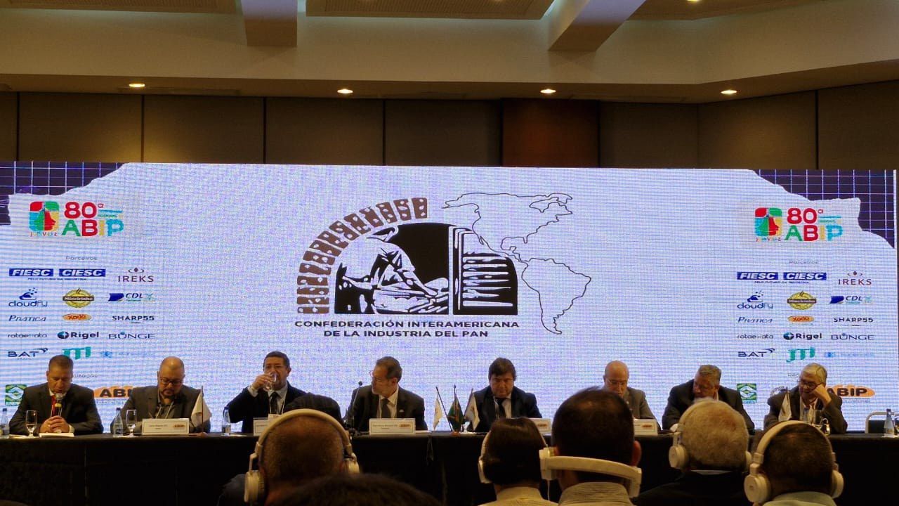 Um grupo de pessoas está sentado em uma mesa em frente a uma tela grande.