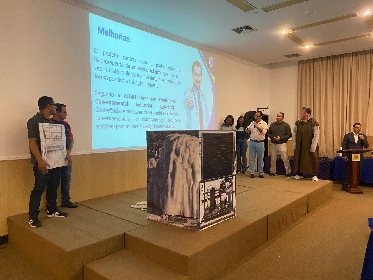 Um grupo de pessoas está em pé em um palco em frente a uma tela de projetor.