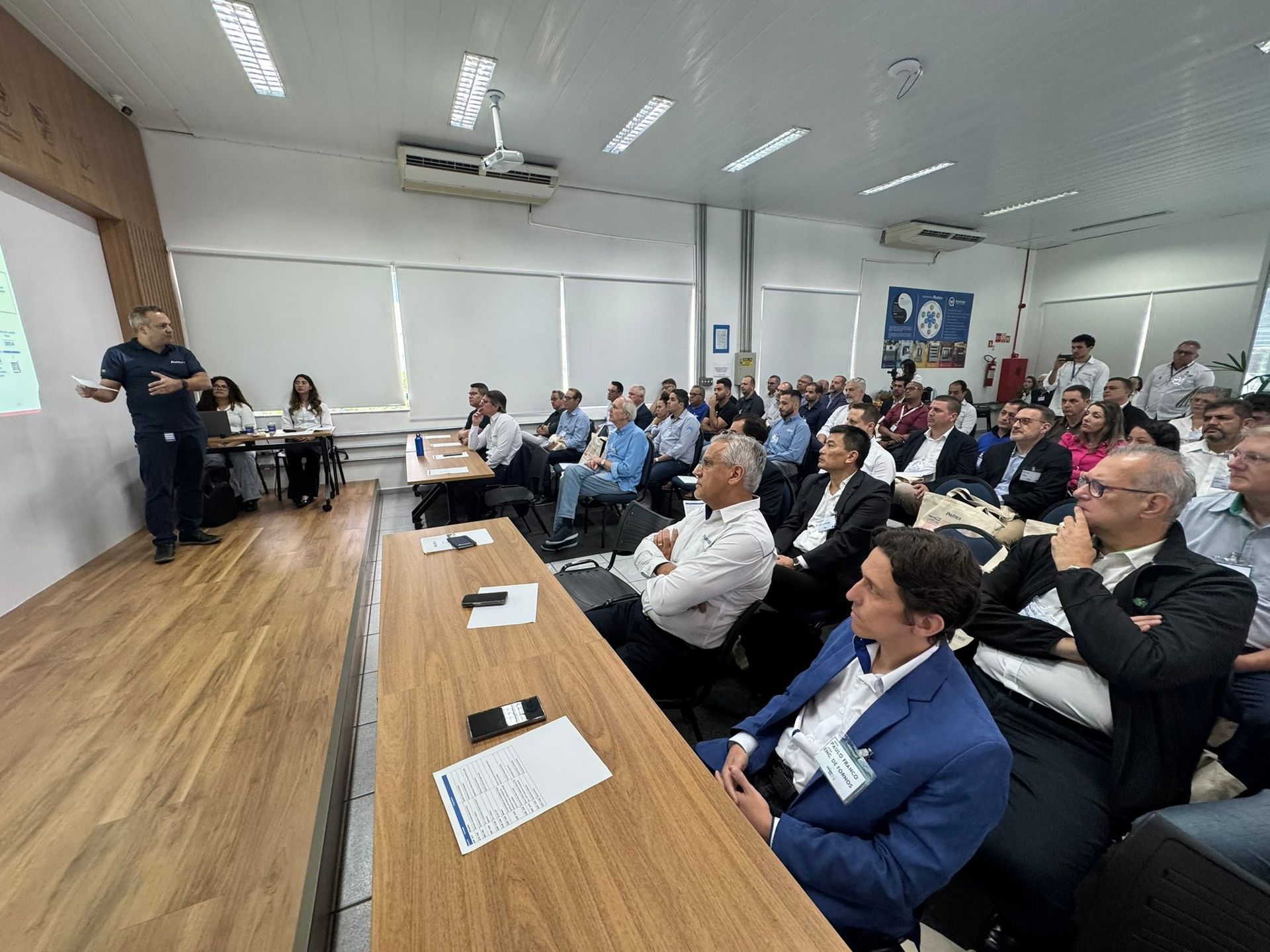 Um grupo de pessoas está sentado em mesas em uma sala assistindo a uma apresentação.