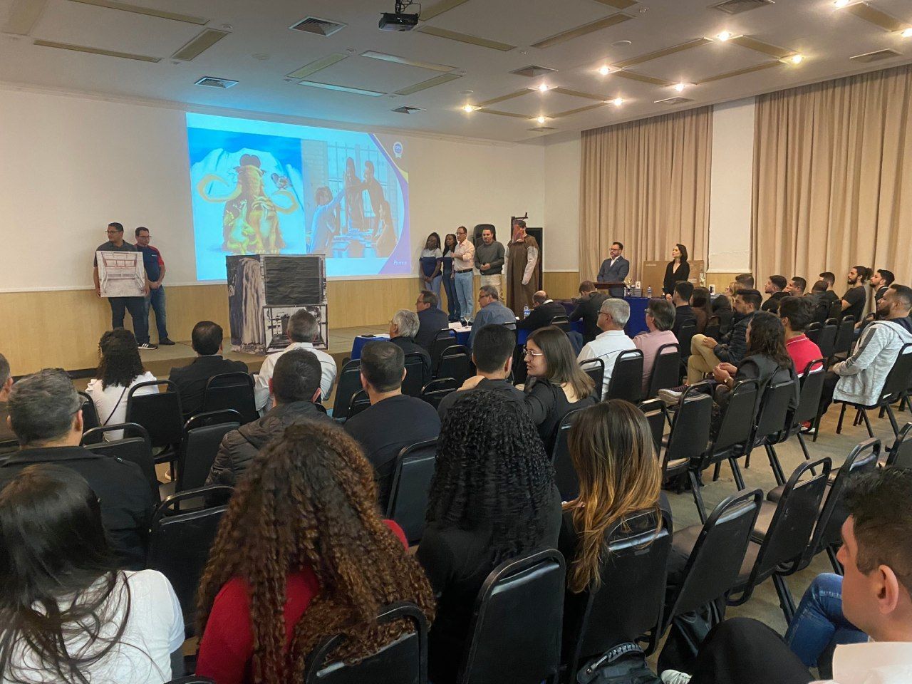 Um grupo de pessoas está sentado em cadeiras em uma sala grande assistindo a uma apresentação.