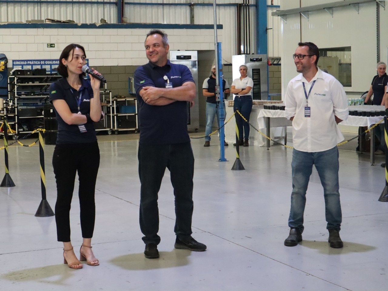 Uma mulher está falando em um microfone enquanto dois homens estão atrás dela
