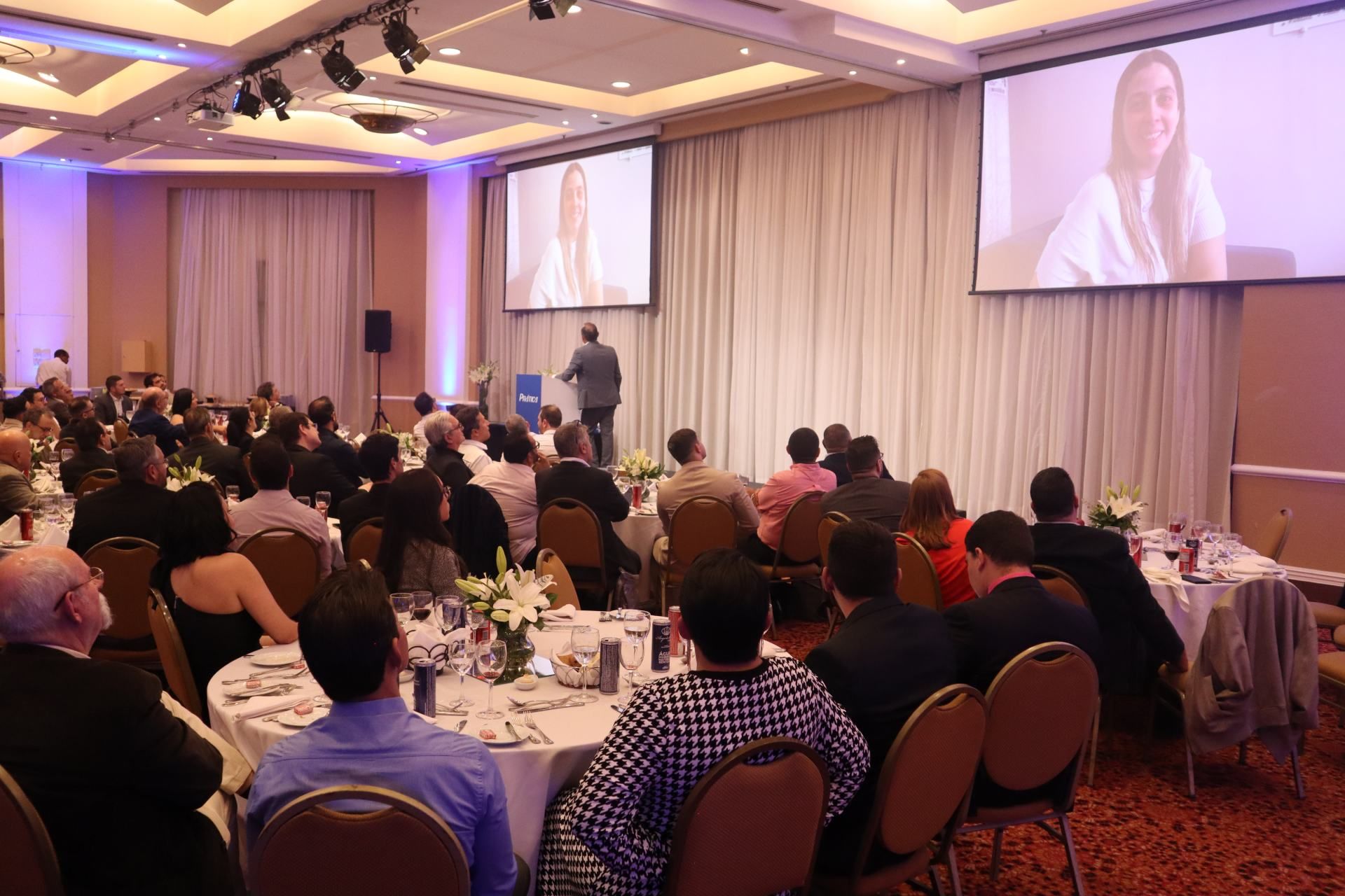 Um grupo de pessoas está sentado em mesas em uma sala grande assistindo a uma apresentação.
