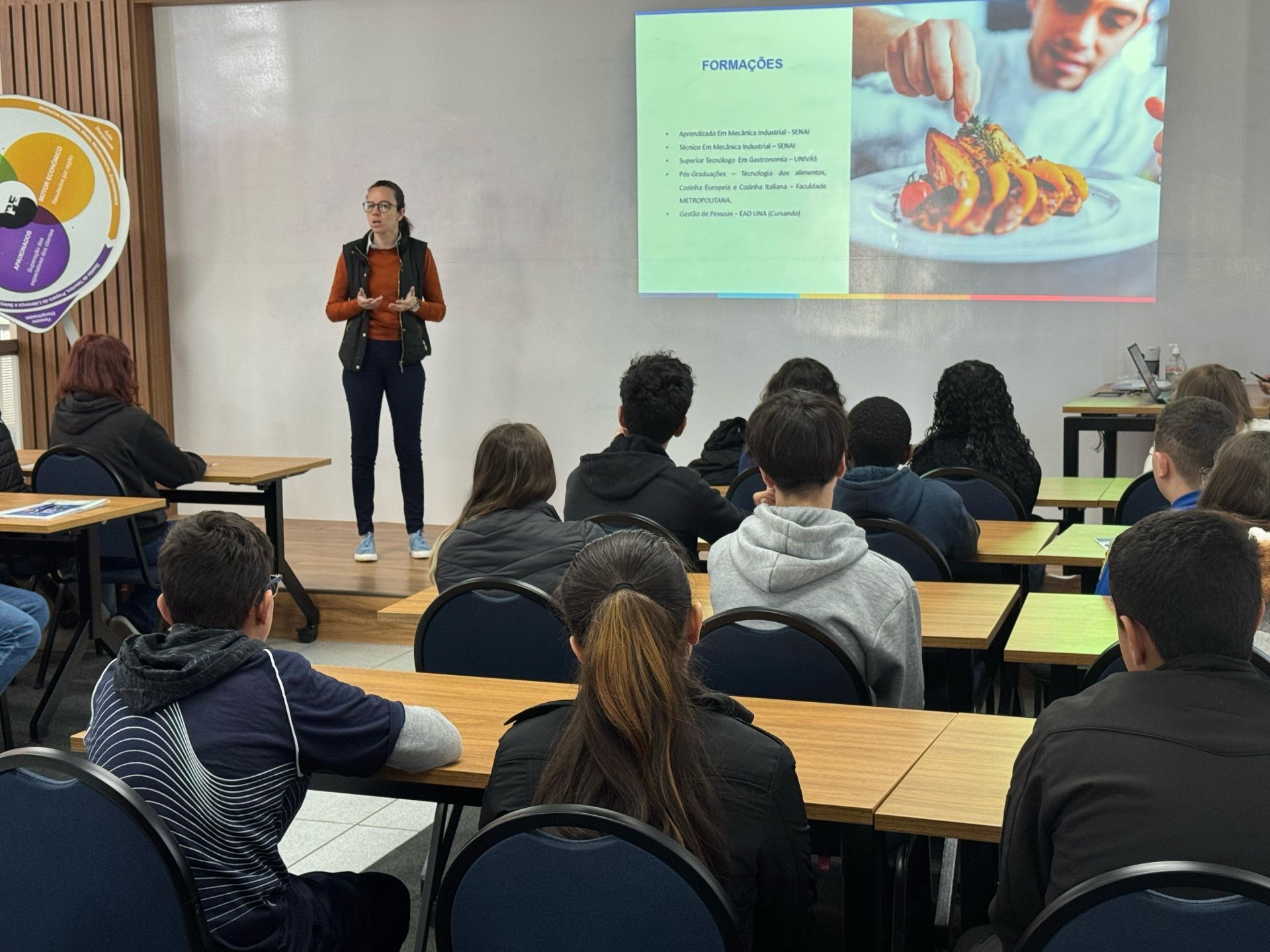 Uma mulher faz uma apresentação sobre comida para uma turma, com uma imagem de tacos em uma tela de projetor.