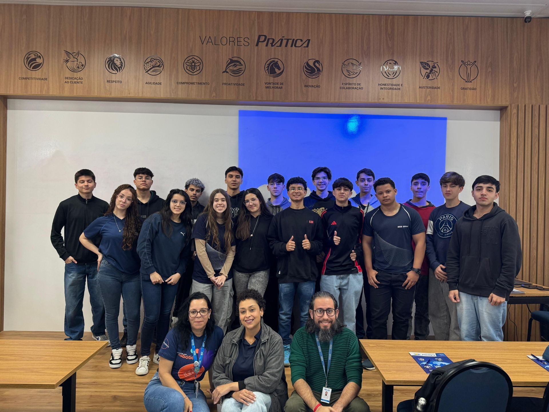 Grupo de estudantes e adultos sorrindo em uma sala de aula; parede de madeira com símbolos; parede com detalhes em azul.