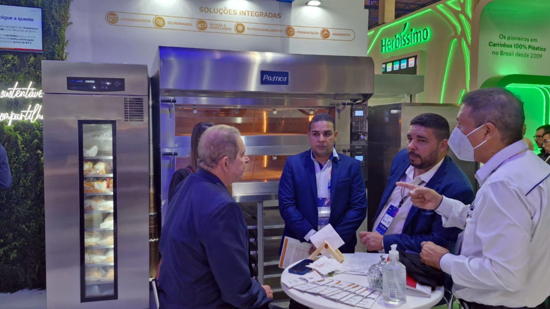 Um grupo de homens está em volta de uma mesa em uma padaria conversando entre si.