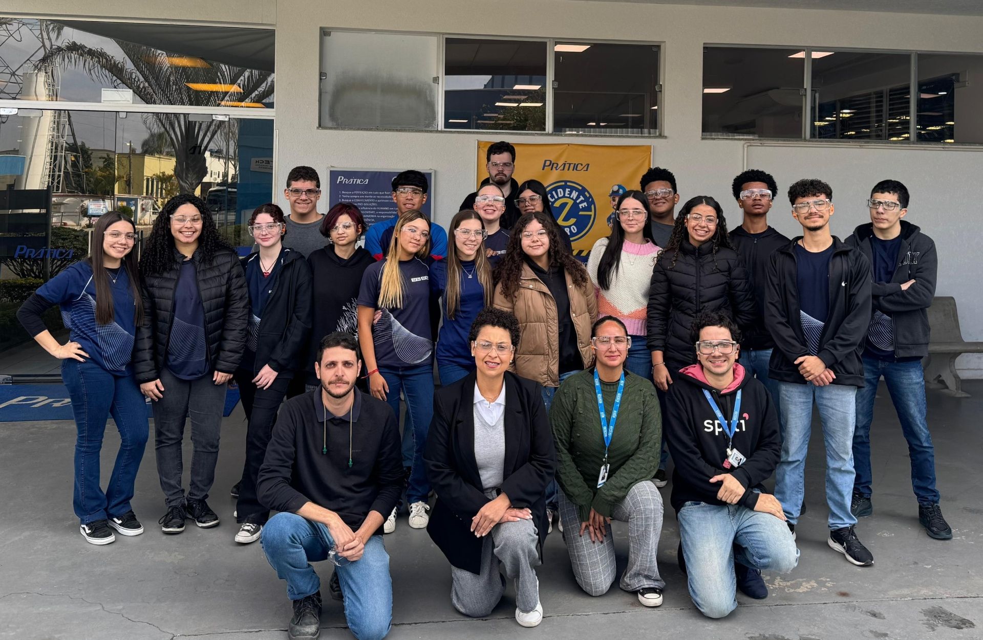 Grupo de pessoas diversas em frente a um prédio, sorrindo. Algumas estão ajoelhadas. Faixa ao fundo.
