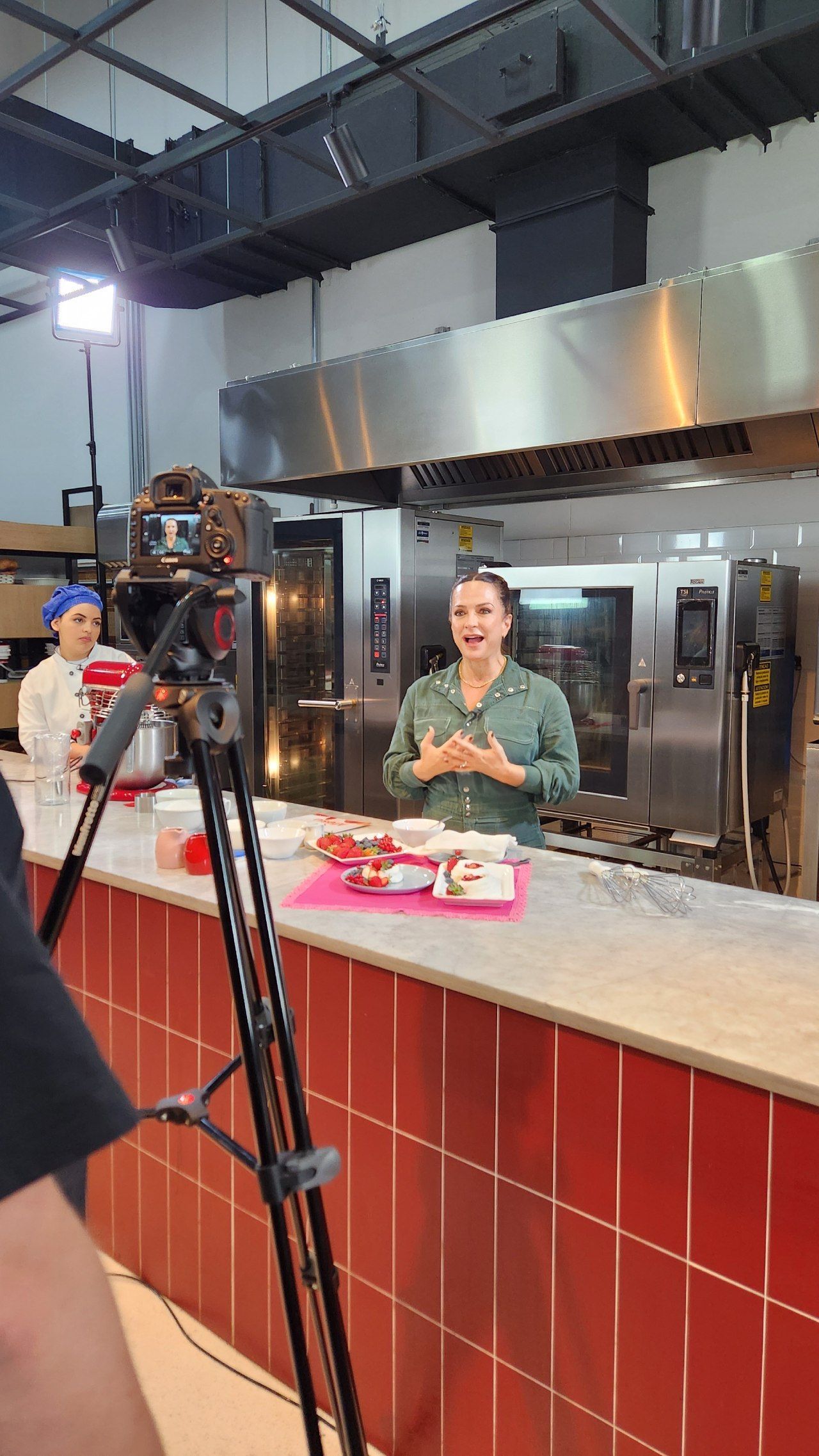 Uma mulher está parada em frente a uma câmera em uma cozinha.