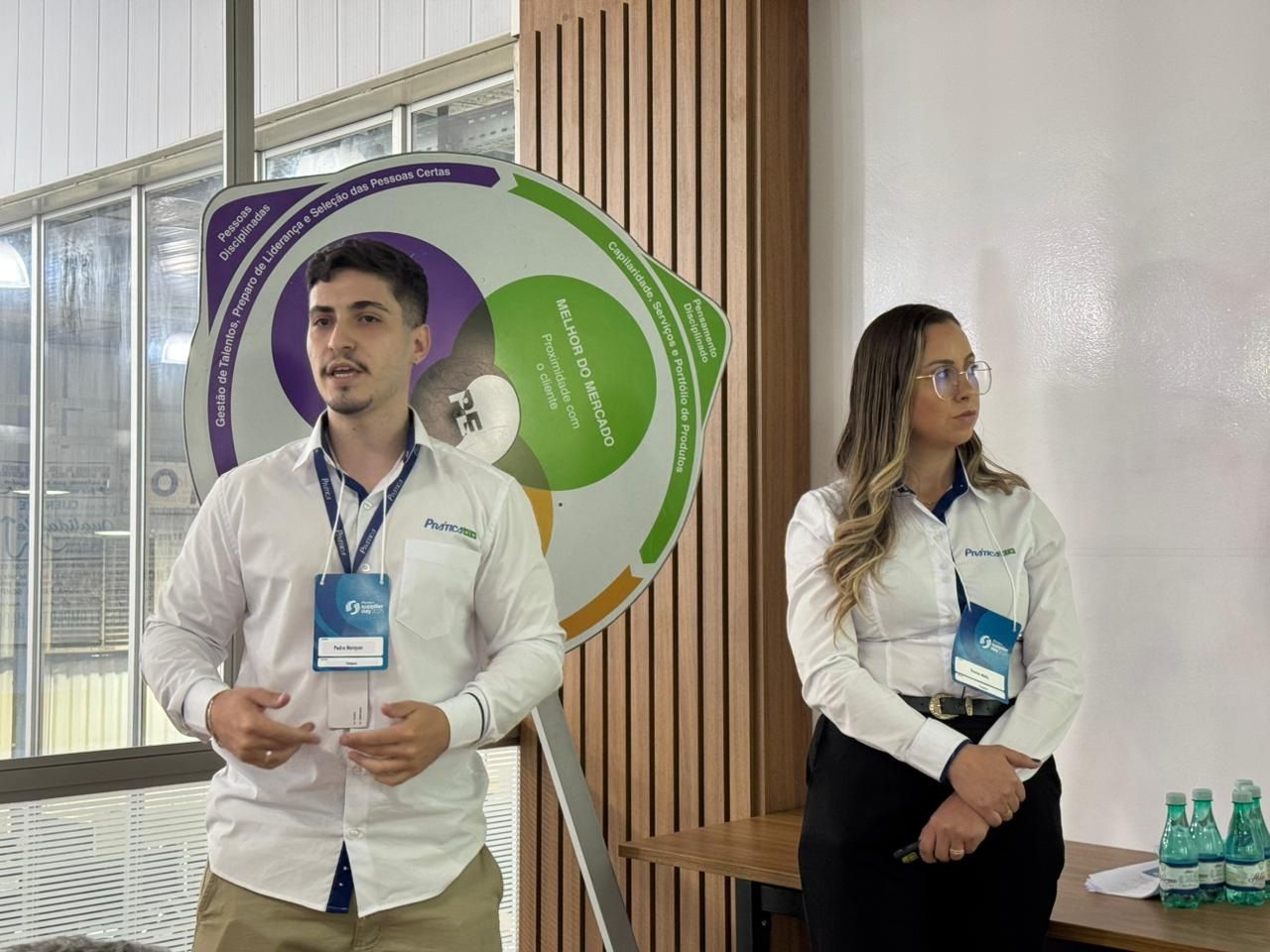 Duas pessoas apresentando: um homem falando e uma mulher ouvindo, em frente a um diagrama colorido.