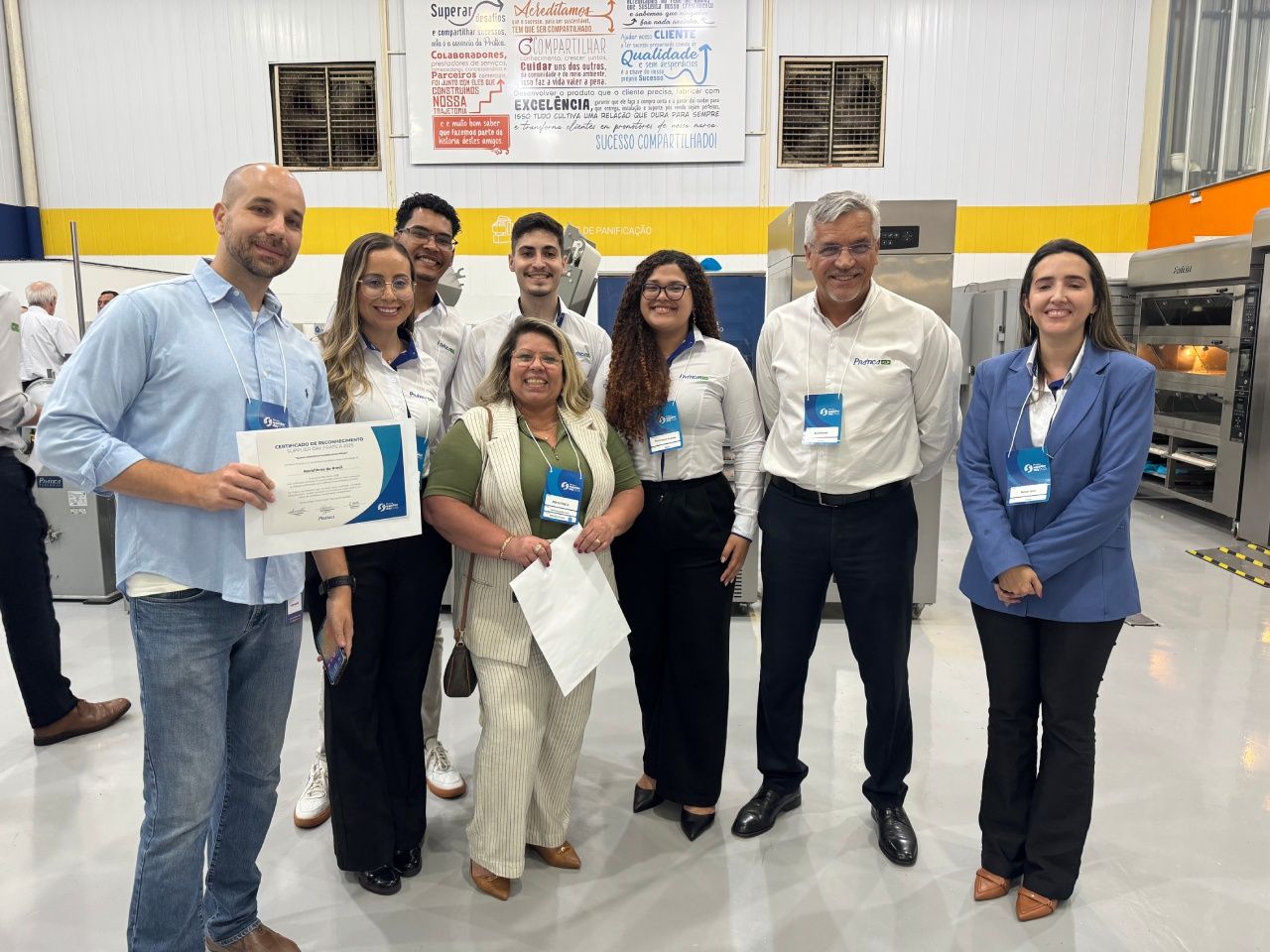 Grupo de pessoas, a maioria em trajes profissionais, posam juntas. Um homem segura um certificado. Ambiente interno.