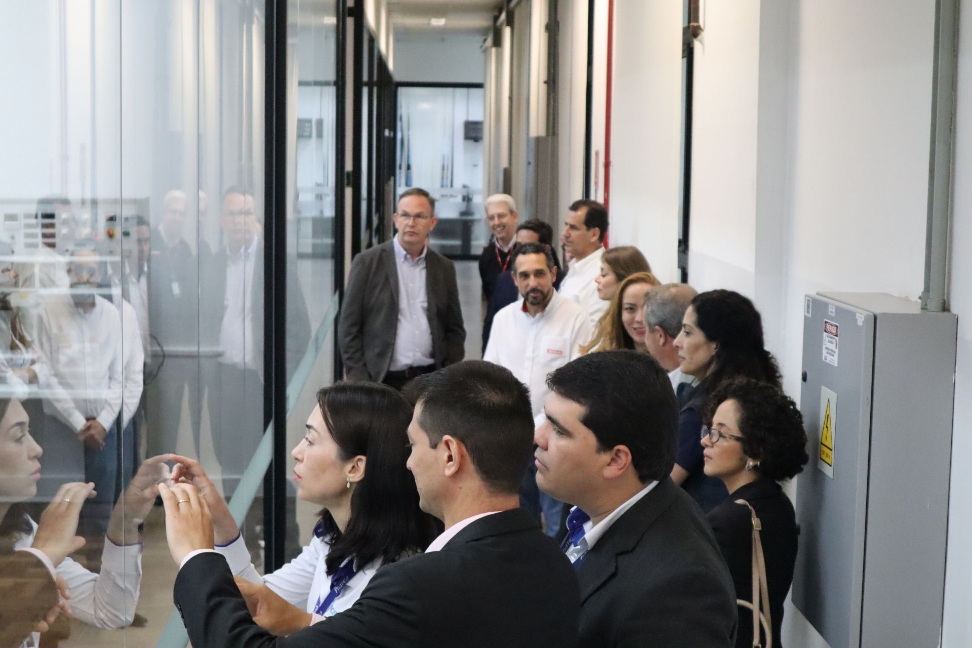 Pessoas olhando através de uma parede de vidro para dentro de uma sala, algumas tirando fotos. Interior, ambiente moderno.