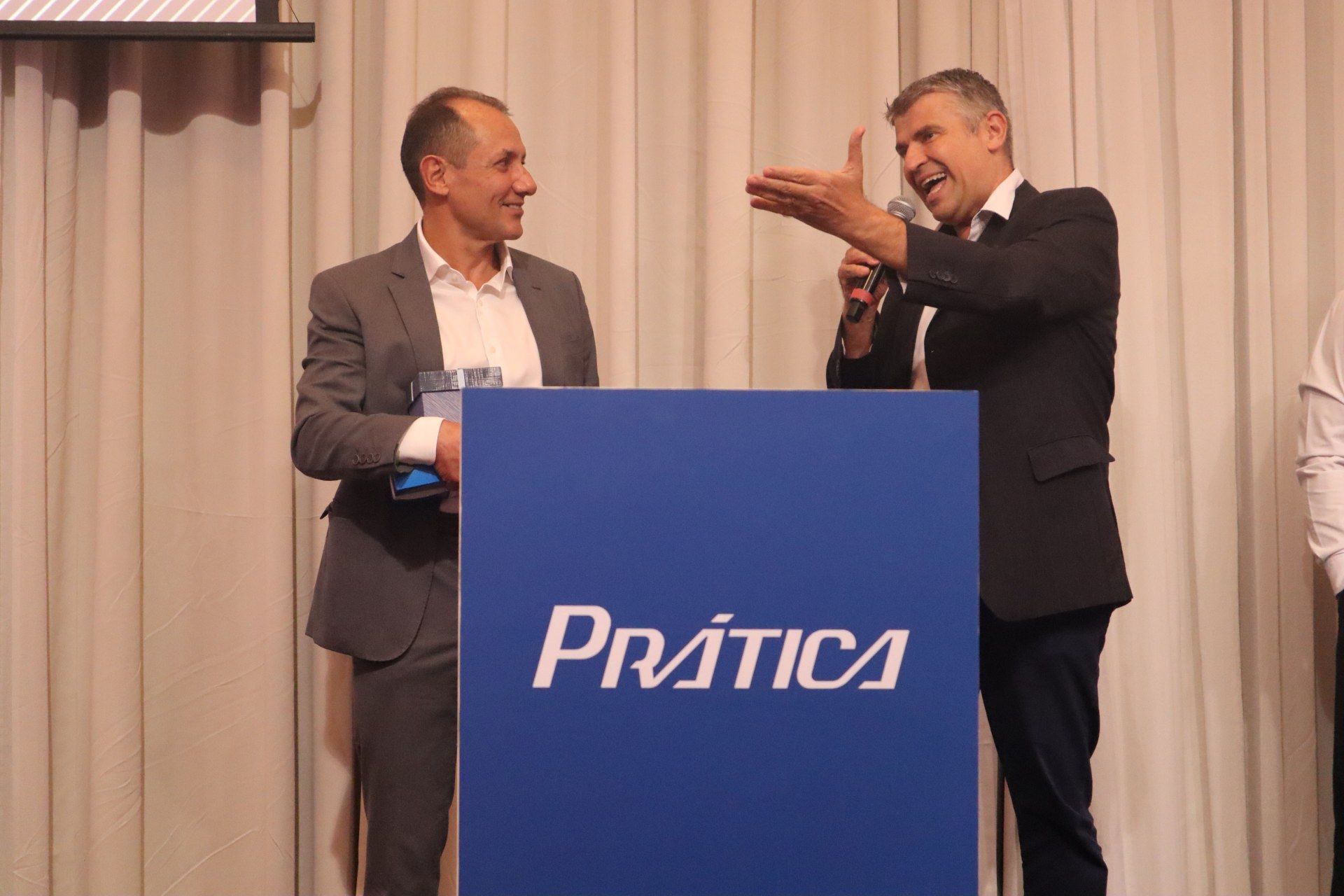 Dois homens estão de pé, um ao lado do outro, em frente a um pódio com a palavra pratica escrita nele.