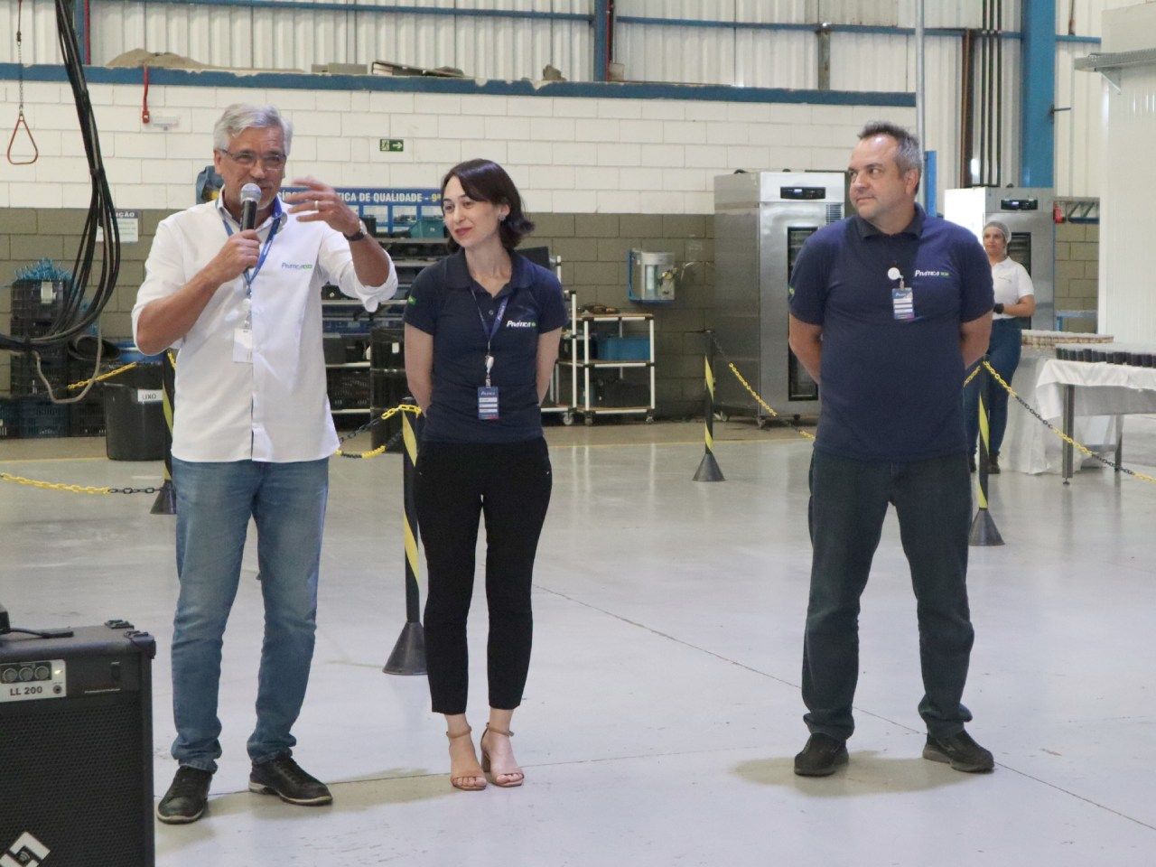 Um homem está falando em um microfone enquanto duas outras pessoas estão atrás dele