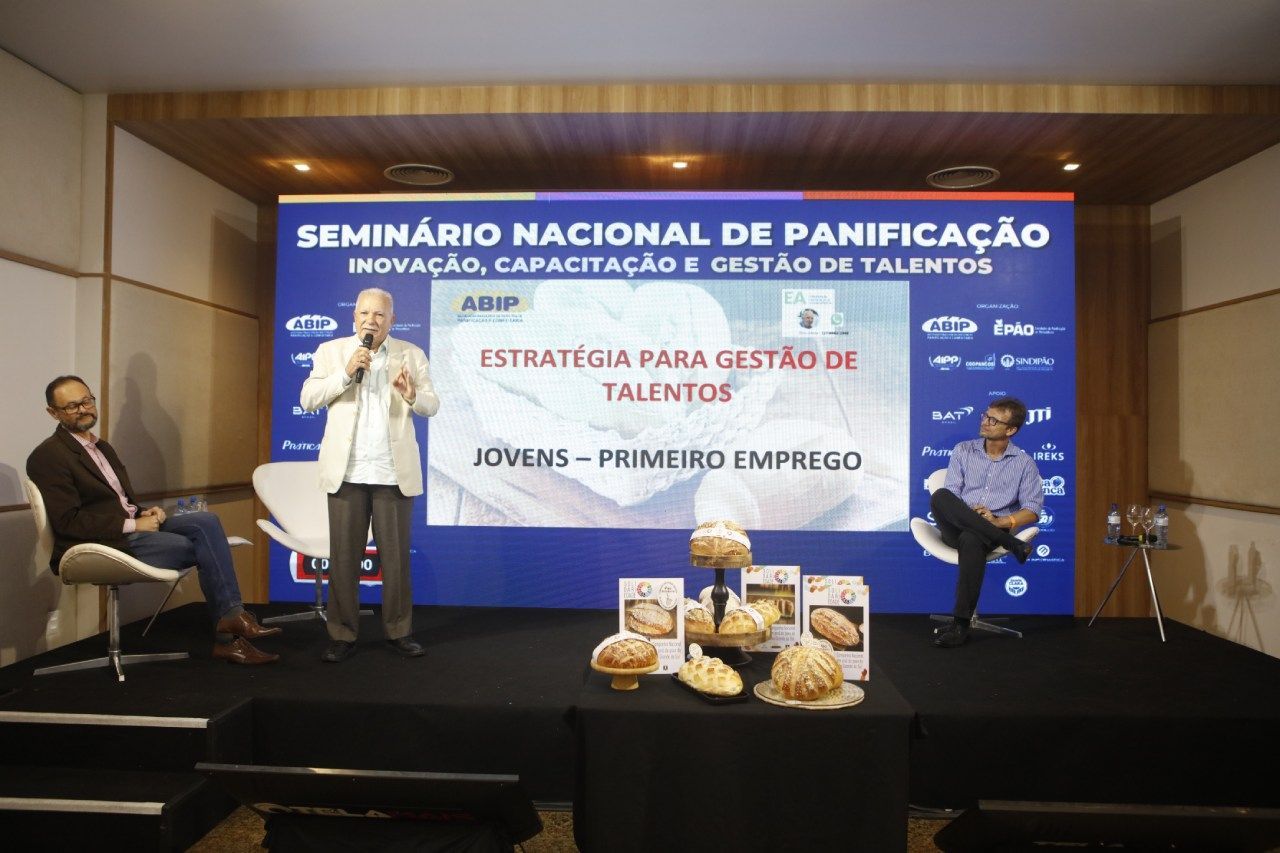 Dois homens estão sentados em cadeiras em frente a uma tela grande.