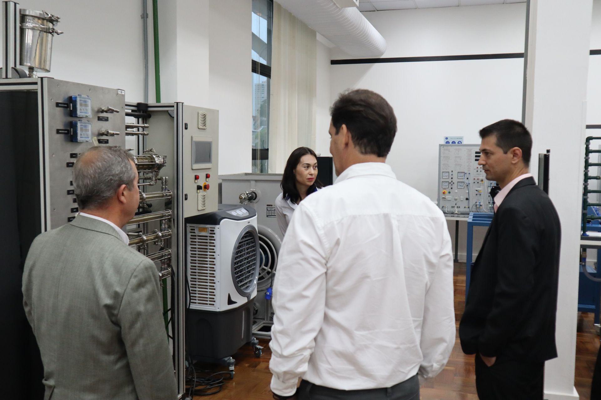 Quatro pessoas em um laboratório, observando equipamentos científicos. Jalecos brancos, diversas máquinas.